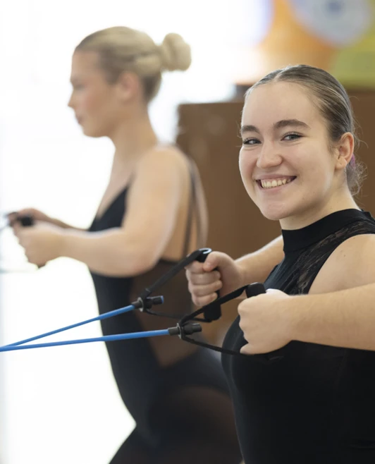 Two dance students using resistance bands, with one smiling confidently during the fitness exercise. Two dance students using resistance bands, with one smiling confidently during the fitness exercise.